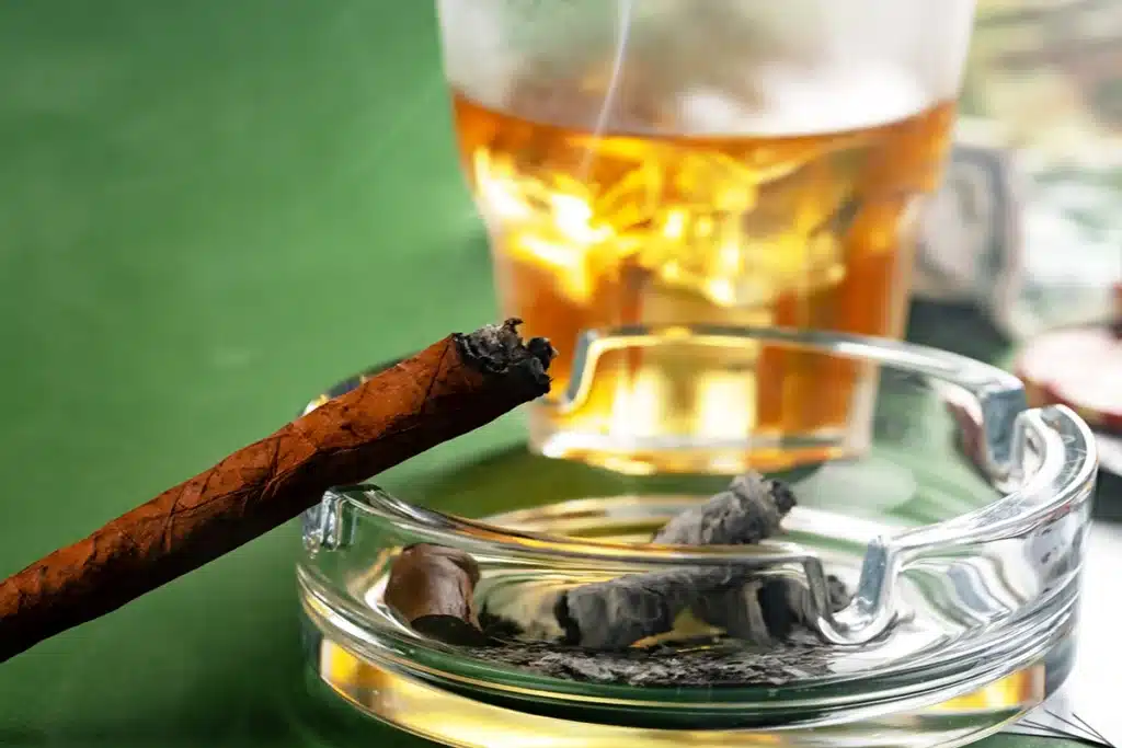 Lit cigar with ash in a tray on a green table with a glass of draft beer in the background